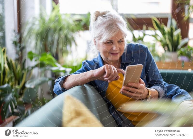 Lächelnde ältere Frau, die ein Smartphone benutzt, während sie zu Hause auf dem Sofa sitzt Menschen lässig Tag Porträt im Innenbereich echte Menschen