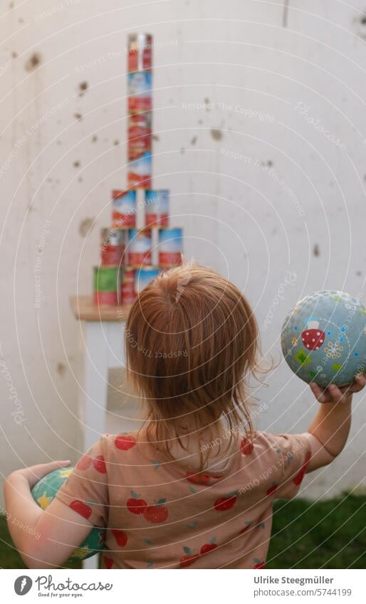 Ein Kind hält zwei Bälle und zielt mit einem Ball auf aufgetürmte Dosen. Das Kind spielt Dosenwerfen. Kindergeburtstag Frühling Sommer Garten Kinderspiel Freude