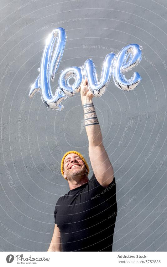 Fröhlicher Mann hält Liebesballon-Briefe Luftballon Freude Glück Beanie gelb grauer Hintergrund Feier Zuneigung Beteiligung glänzend metallisch Zauber Emotion