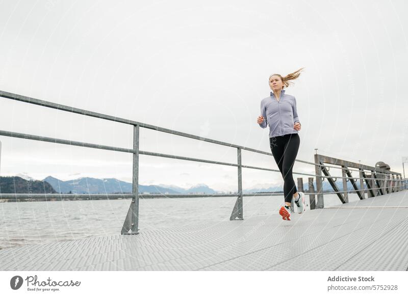 Junge Frau joggt entlang einer ruhigen Uferpromenade Joggen Hafengebiet Athlet laufen Promenade Bucht Berge u. Gebirge Hintergrund jung im Freien Übung Fitness