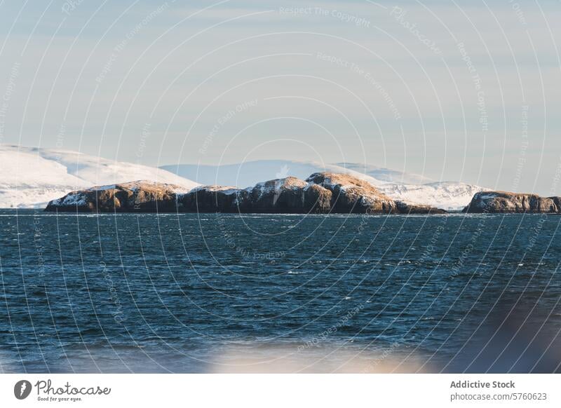 Schneebedeckte Felseninseln tauchen aus dem eisblauen Wasser vor der isländischen Küste auf, umrahmt von einem klaren Himmel und fernen Schneegipfeln Insel