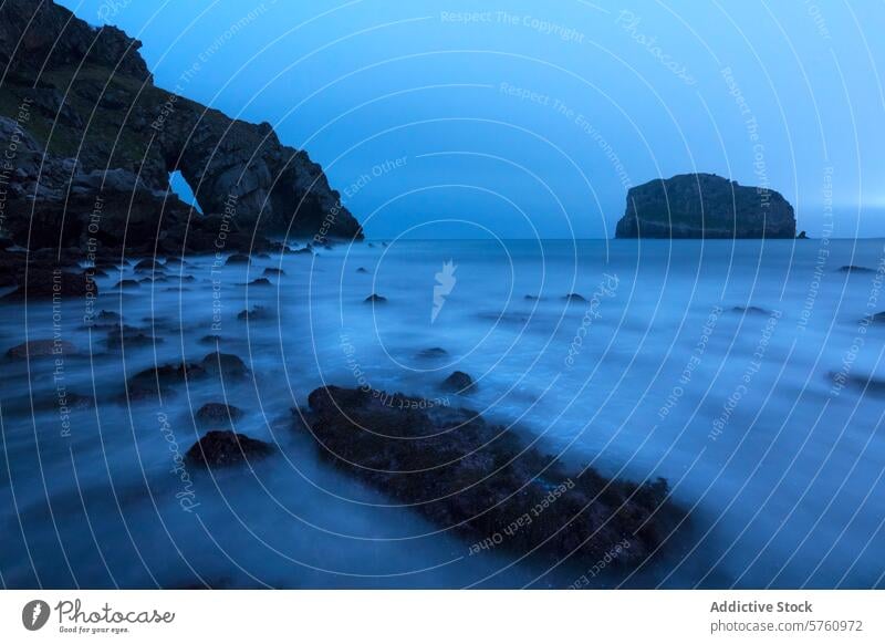 Die heitere blaue Stunde bricht über den ikonischen San Juan de Gaztelugatxe herein, mit seinem natürlichen Bogen und dem felsigen Ufer, das sanft im Meer verschwimmt.