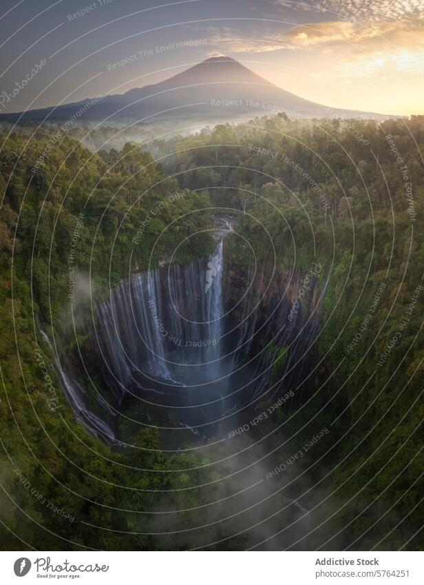 Das erste Licht der Morgendämmerung beleuchtet einen nebligen, majestätischen Wasserfall mit einem hoch aufragenden Vulkan in der Ferne, inmitten des tropischen Waldes von Indonesien