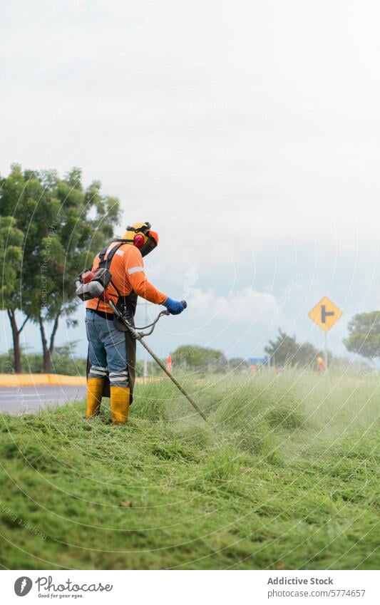 Landschaftsgärtner beim Schneiden von Gras mit einer Unkrautsense Trimmen Sense Sicherheit Ausrüstung Straßenrand Gehör Schutz Uniform orange hoch Arbeit