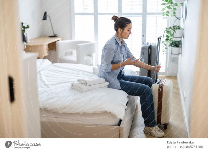 Junge Frau mit einem Koffer sitzt auf dem Bett in einem Hotelzimmer und telefoniert Verpackung Bekleidung Ferien reisen Vorbereitung Menschen Gepäck eine Person