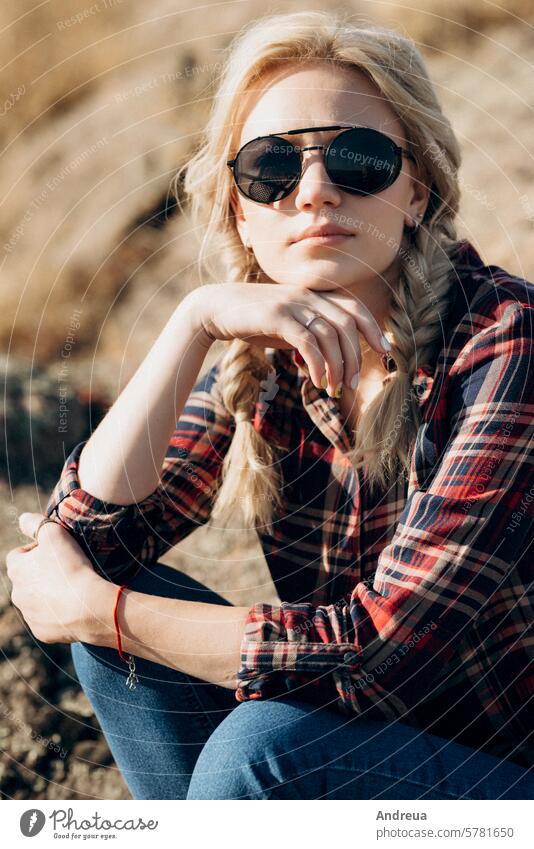 blondes Mädchen mit Zöpfen in einem Hemd, Jeans, roten Schuhen und schwarzer Brille vor dem Hintergrund Herbst blau Stiefel braun modisch Granit Gras Frisur