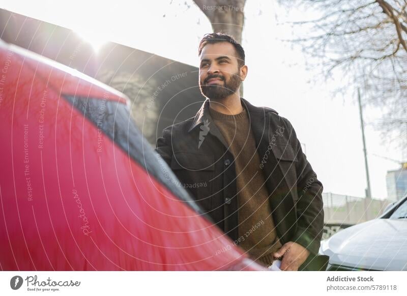 Ein eleganter Mann steht neben einem roten Auto und blickt nachdenklich in die Ferne, gebadet im Sonnenlicht, mit Stadtbäumen im Hintergrund. PKW selbstbewusst