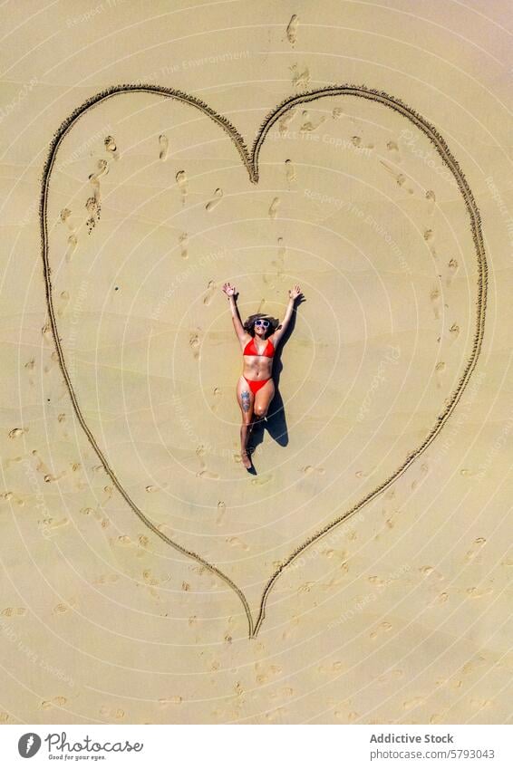 Fröhlicher Strandtag mit einem Herz im Sand Frau Luftaufnahme sonnig Seeküste liegend Sandzeichnung Uferlinie Badebekleidung Urlaub Feiertag Sonnenbrille Sommer