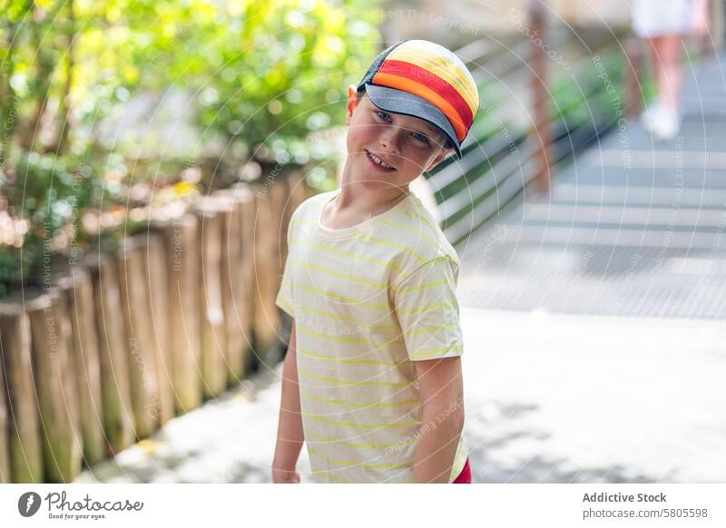 Junger Junge mit bunter Mütze lächelt im Freien Lächeln Verschlussdeckel gestreift gelb T-Shirt heiter Sommer Park Natur Kind jung Glück Sonnenlicht Tag lässig