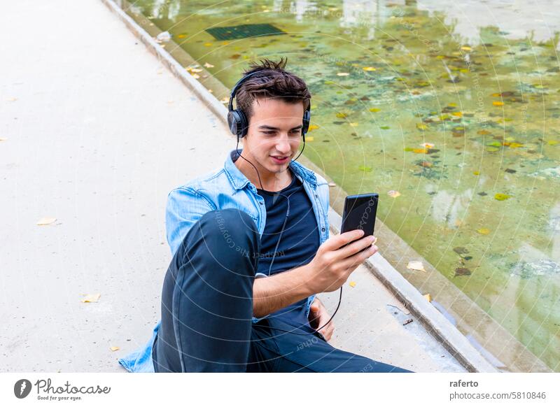 Mann hört Musik an einem Teich. Kopfhörer Smartphone hören Jeansstoff Hemd schwarz Hose jung Erwachsener fokussiert entspannt urban im Freien Sitzen lässig