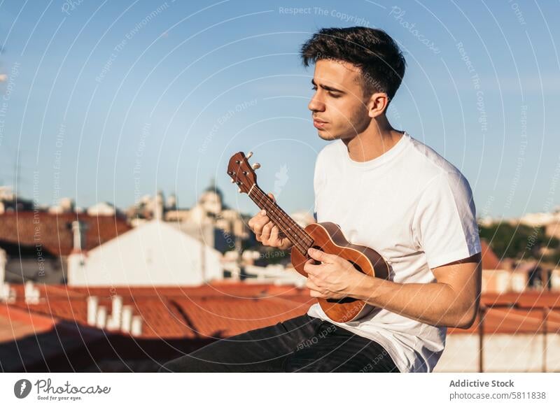 junger Mann sitzend und Ukulele spielend auf einem Dach Person Musik Gitarre Instrument Musical Musiker akustisch Hintergrund Erwachsener Spaß Glück männlich