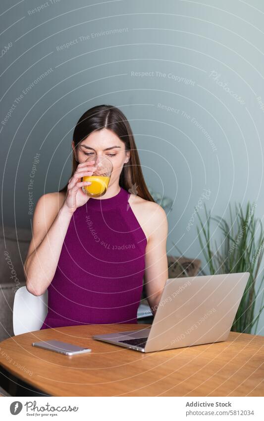 Fröhliche Frau, die im Wohnzimmer am Laptop sitzt benutzend Zahnfarbenes Lächeln Glück Apparatur Browsen Saft trinken heiter Netbook lässig Glas orange