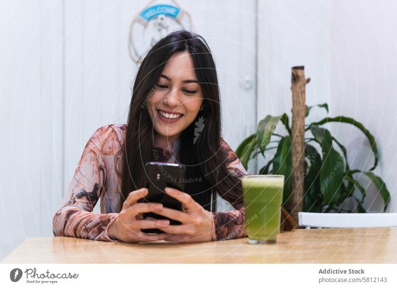 Lächelnde Frau mit Smartphone am Tisch in einer Bar benutzend Kälte Smoothie Erfrischung trinken Browsen Getränk Mobile Gerät Internet ruhen Glück Apparatur