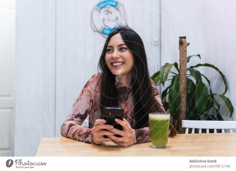 Lächelnde Frau mit Smartphone am Tisch in einer Bar benutzend Kälte Smoothie Erfrischung trinken Browsen Getränk Mobile Gerät Internet ruhen Glück Apparatur