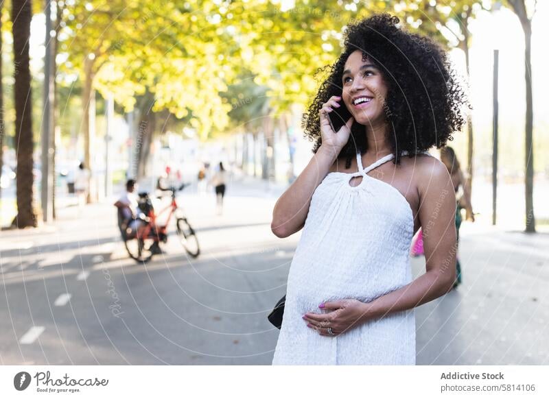 Lächelnde schwarze schwangere Frau, die mit ihrem Smartphone spricht benutzend reden Gespräch positiv Bauch Talkrunde warten sprechen Funktelefon Gerät