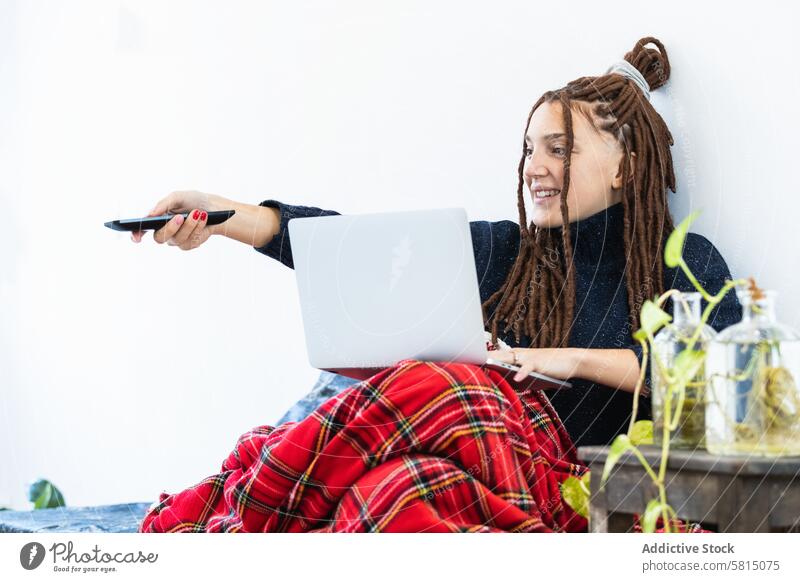 Frau mit Dreadlocks, die zu Hause an einem Laptop arbeitet und eine Fernbedienung in der Hand hat heimwärts Computer Sofa Sitzen Raum Internet