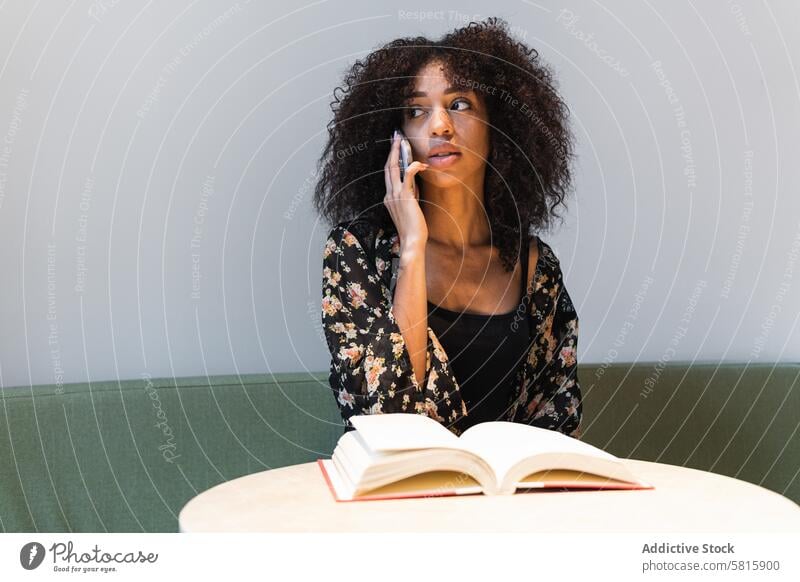 Ethnische Frau, die am Tisch mit einem Buch über ihr Smartphone spricht reden feminin nachdenken Afro-Look Frisur Sofa benutzend Apparatur Gerät verträumt