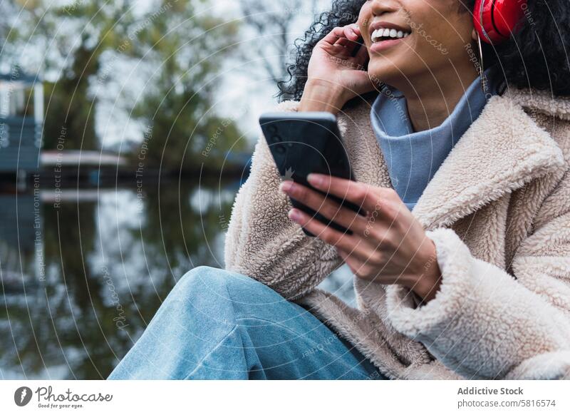 Fröhliche schwarze Frau beim Musikhören im Park Kopfhörer Smartphone benutzend zuhören Kälte ruhen sich[Akk] entspannen See afroamerikanische Frau Großstadt