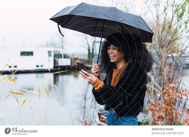 Fröhliche schwarze Frau mit Smartphone und Regenschirm Großstadt benutzend Straße urban Vorschein Mobile Afroamerikaner jung Glück positiv Lächeln heiter Saison