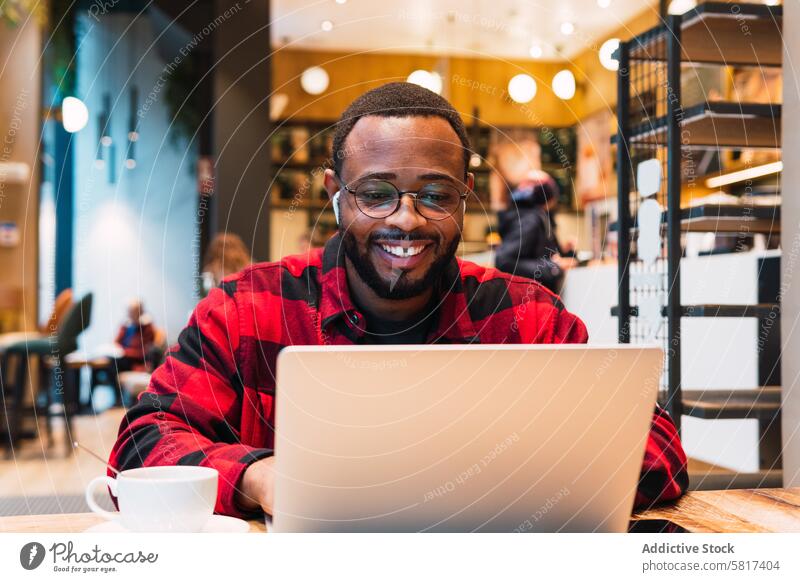 Lächelnder schwarzer Mann arbeitet an einem Laptop in einem Café Kopfhörer benutzend online Internet Browsen freiberuflich Arbeit männlich Afroamerikaner