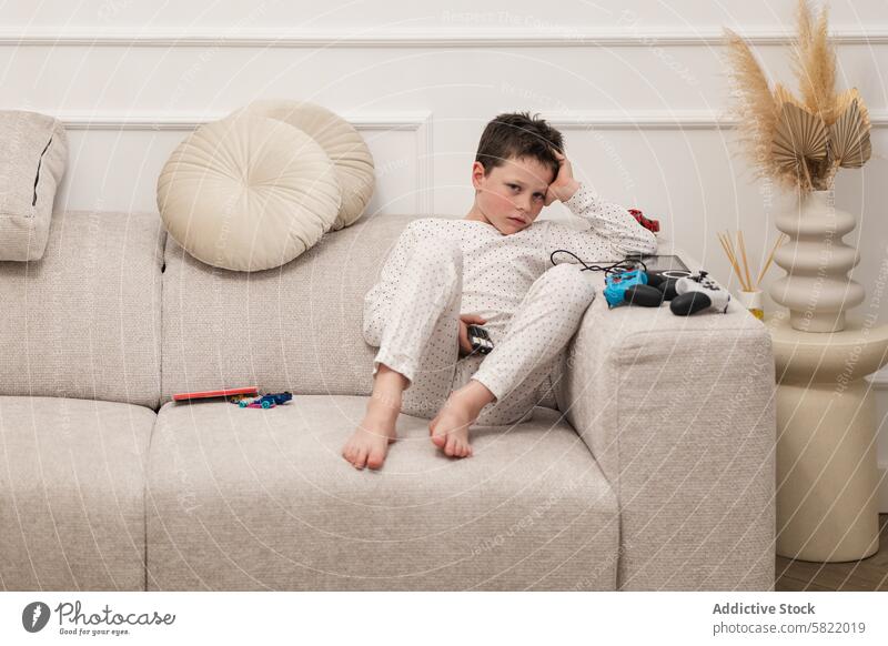 Junge mit Spielzeug und Spielcontroller auf der Couch liegend heimwärts Liege faulenzen Pyjama gelangweilt Game-Controller Wegsehen beige im Innenbereich Kind