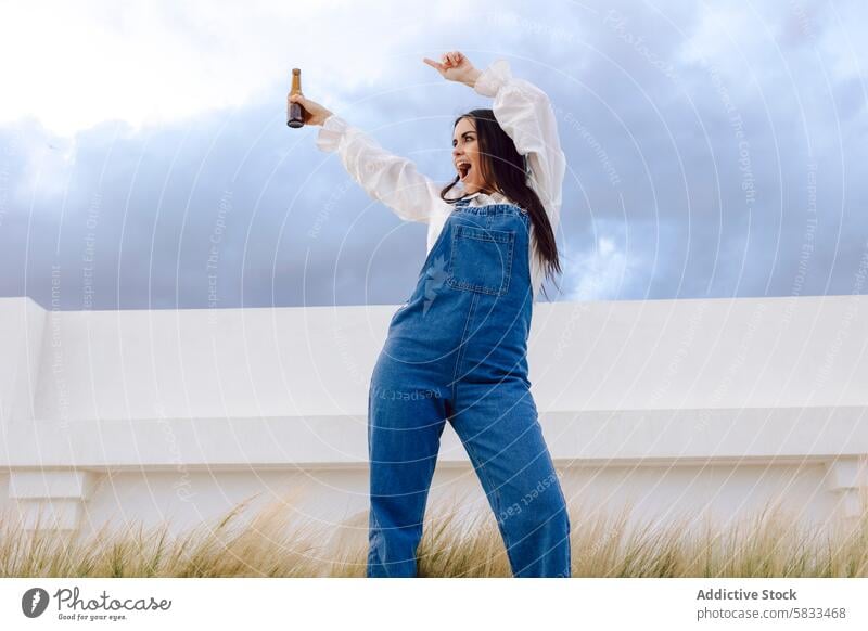 Feiernde Frau mit Bier am Hafen Freude Flasche Jeansstoff Latzhose Portwein Himmel Wolken festlich Pose Beteiligung Alkohol Freizeit Dock Hafengebiet maritim