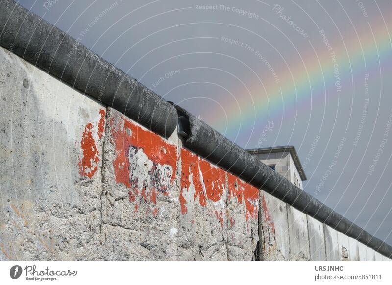 Ausschnitt der Mauer verläuft diagonal, im Hintergrund ragt ein Teil des oberen Stockwerks eines Gebäudes hervor, ein bunter Regenbogen von West nach Ost verbindet wie eine Brücke