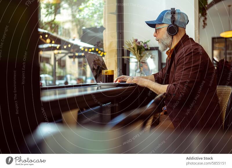 Moderner älterer Mann, der aus der Ferne in einem Café arbeitet Senior modern Fernarbeit Lifestyle Laptop Kopfhörer Technik & Technologie Baseballmütze stylisch