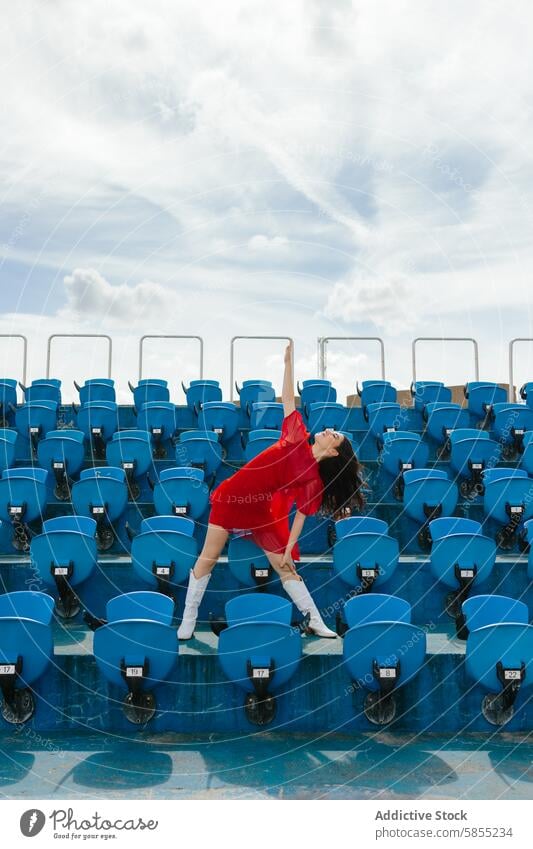 Junge Frau tanzt freudig in einem Stadion Tanzen Sitz rot Kleid blau Himmel Cloud Freude Energie jugendlich Bewegung im Freien Feier Mode Stil Glück pulsierend