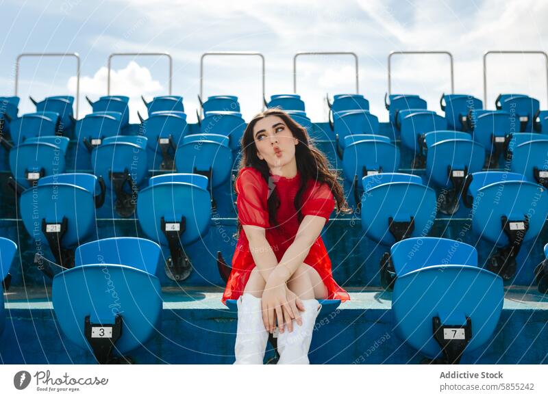 Junge Frau allein in einem Stadion, die einen Kuss sendet Sitz rot blau spielerisch im Freien Himmel Tag Sommer jung lässig Mode Stil urban Sitzgelegenheit leer