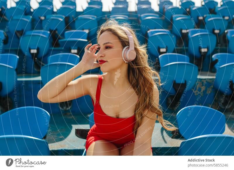 Junge Frau genießt Musik auf einem Sitzplatz im Stadion Kopfhörer Sitzgelegenheit sonnig Tag rot Badeanzug blau hören jung Genuss Freizeit im Freien