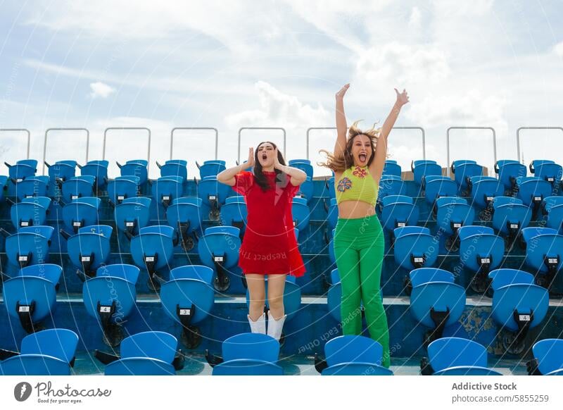 Junge Frauen, die in einem Stadion enthusiastisch jubeln jauchzen springen energetisch leer Sitz blau Himmel pulsierend Outfit Freund Freude Aufregung Feier