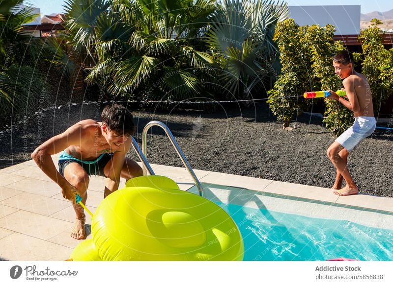 Verspielte Geschwister bei einer Wasserpistolenschlacht am Pool Spaß Junge Geschwisterkind spielen Ente zielen Badebekleidung im Freien Sommer Aktivität Spiel