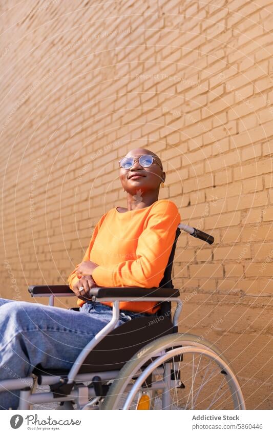 Selbstbewusste Frau im Rollstuhl in der Nähe einer Steinmauer selbstbewusst Afroamerikaner schwarze Frau modern stylisch urban außerhalb sonnig Backsteinwand