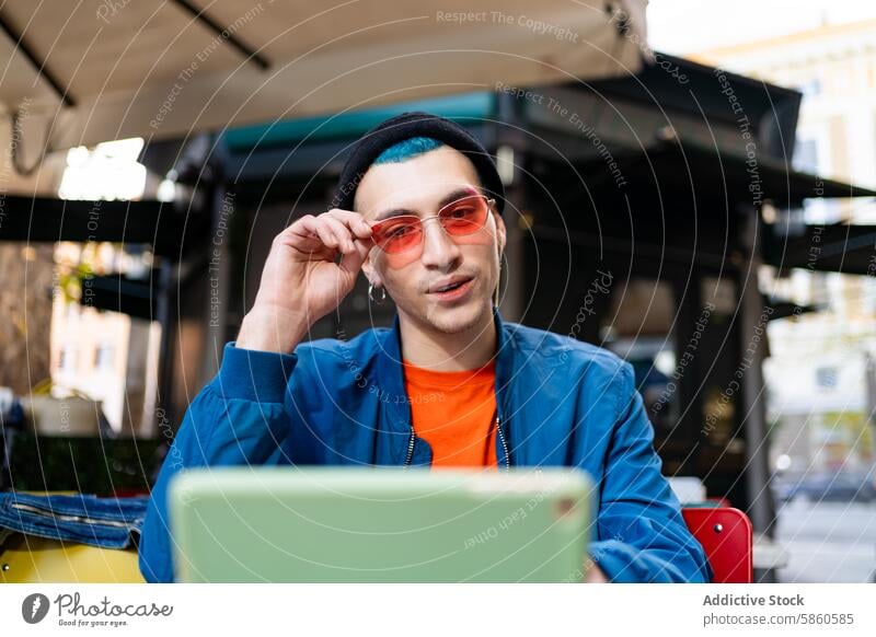 Stilvoller junger Mann passt seine Sonnenbrille im Café an stylisch im Freien Laptop Mode urban lässig Sitzen Einstellung berühren rosarot blau Jacke orange