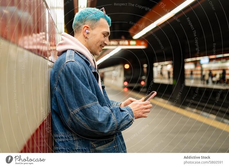 Junger Mann mit blauen Haaren benutzt Smartphone in einer U-Bahn-Station jung blaue Haare Warten Zug Jeansstoff Jacke Im-Ohr-Gerät Kopfhörer Mobile Telefon