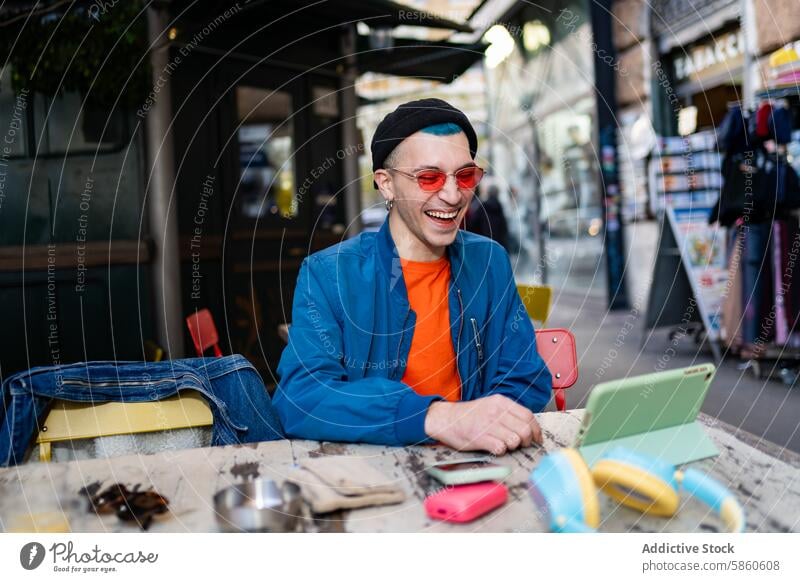 Stylischer junger Mann lachend in einem Café mit Smartphone und Kopfhörern Lachen im Freien Tisch Beanie Sonnenbrille rot blau Jacke Freude Fröhlichkeit