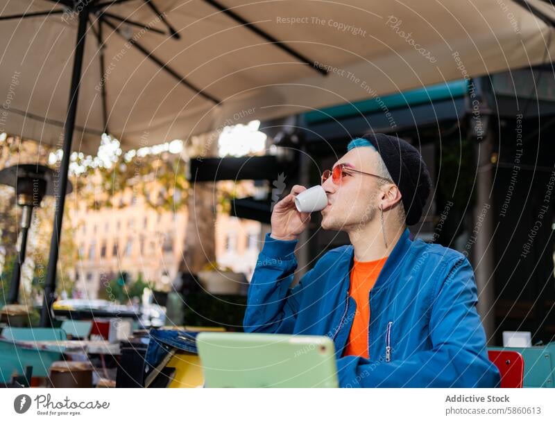 Stilvoller junger Mann genießt einen Kaffee in einem Café im Freien Espresso Straße trendy stylisch Mode blau Jacke Sonnenbrille Beanie schlürfend Sitzen Tisch