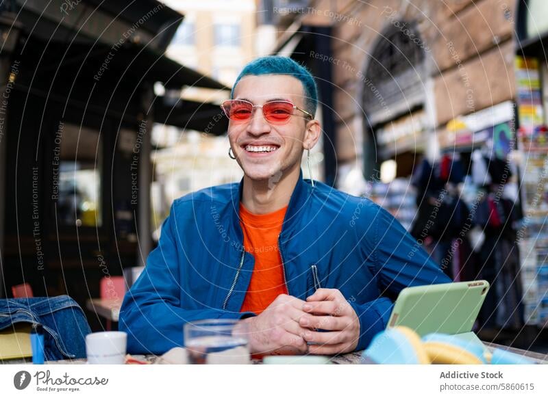 Lächelnder junger Mann mit blauen Haaren genießt einen Kaffee im Freien blaue Haare rote Sonnenbrille Café Smartphone Tisch Fröhlichkeit Freude Mode Stil urban