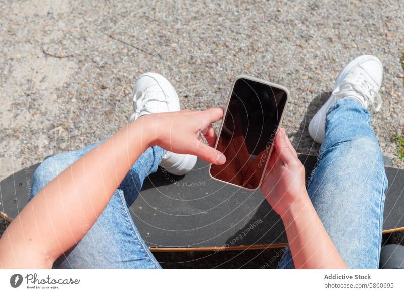 An einem sonnigen Tag auf dem Skateboard sitzend und das Smartphone benutzend Person Jeanshose Turnschuh im Freien lässig Technik & Technologie Straßenbelag