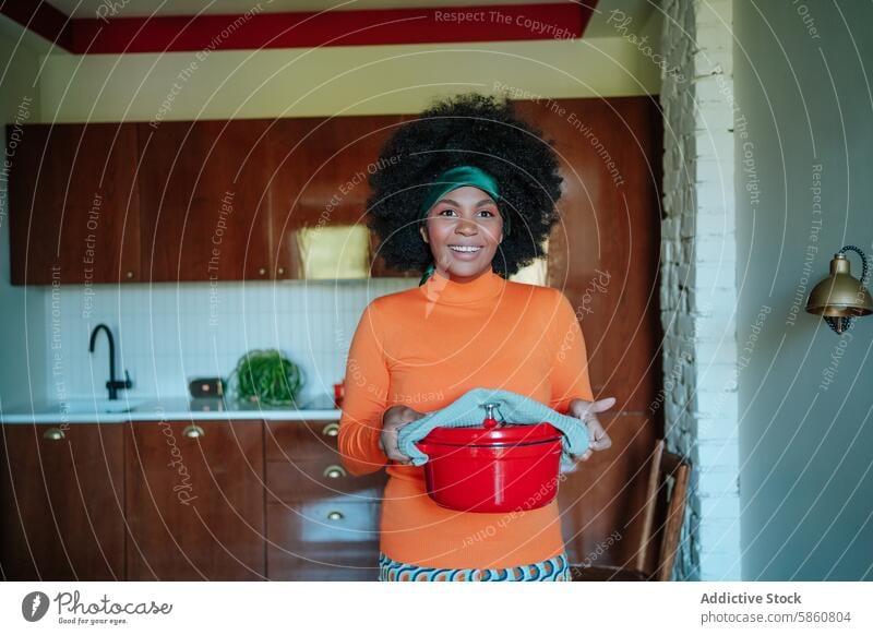 Lächelnde Frau in einer Retro-Küche, die einen roten Topf hält schwarz retro 1950s Essen zubereiten in die Kamera schauen heiter orange Kleid pulsierend