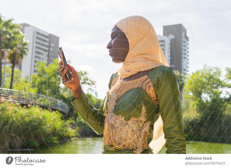 Frau in traditioneller Kleidung telefoniert in einem Stadtpark Park Smartphone Hijab urban Grün Gebäude sonnenbeschienen Großstadt Natur im Freien