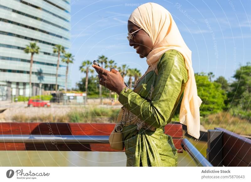 Frau mit Hidschab, die lächelnd ihr Telefon im Freien benutzt Hijab Smartphone sonnig heiter modern Gebäude Handfläche Baum grün pulsierend muslimisch genießend