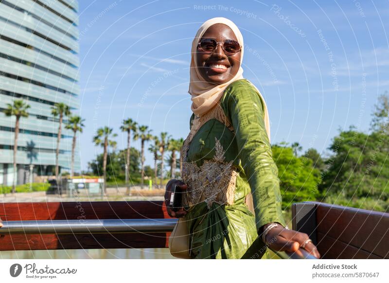 Lächelnde Frau im Hidschab genießt den sonnigen Park Hijab grün Kleid bestickt urban Tag friedlich freudige Bank Palme im Freien Freizeit Fröhlichkeit Mode Baum