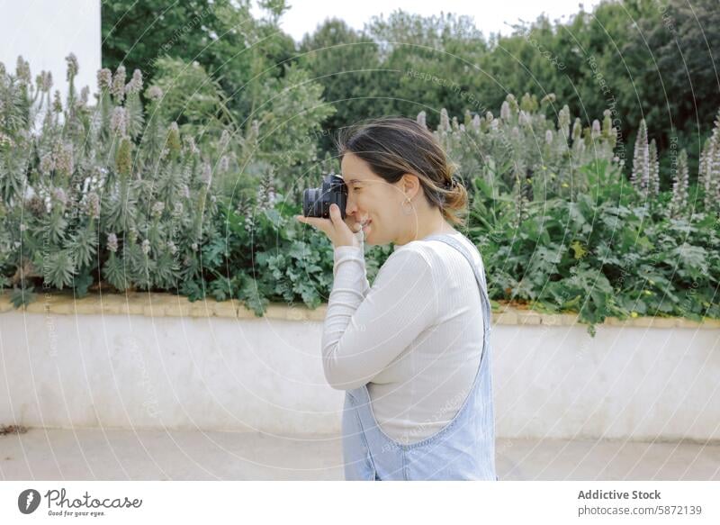 Frau in Freizeitkleidung fotografiert Gartenpflanzen Fotokamera Fotografie Pflanzen im Freien Jeansstoff Overall weißes Hemd Natur Laubwerk Hobby tagsüber