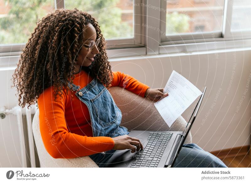 Frau erledigt Papierkram und benutzt Laptop im Coworking Space Afroamerikaner Schriftstück Arbeit Fernarbeit lässig Brille krause Haare Jeansstoff