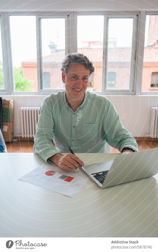 Lächelnder Geschäftsmann in einem hellen Büro, der an einem Laptop arbeitet Arbeit Fenster urban Gebäude Hemd grün Tisch Papier Schreibstift Sitzen Ansicht