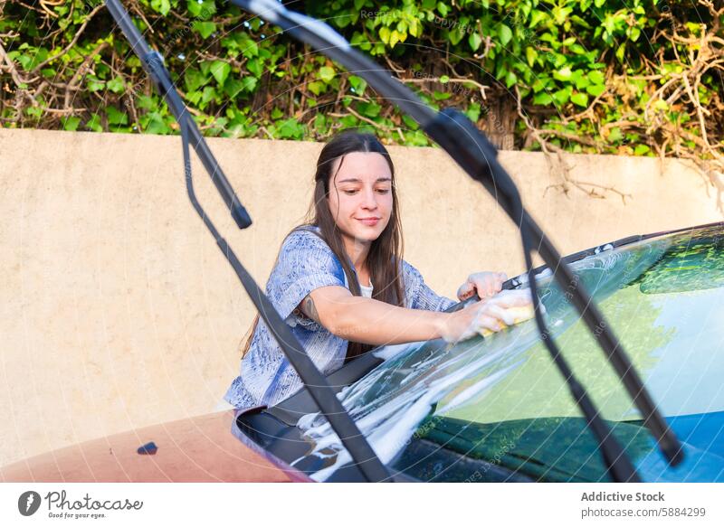 Junge Frau wäscht ihr Auto an einem sonnigen Tag PKW Waschen Wäsche waschen Autowäsche Fahrzeug Reinigen Windschutzscheibe Schwamm Seife Aufgabe im Freien