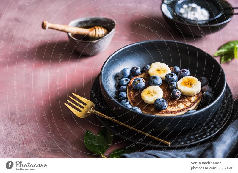 Pancake mit Bananen, Heidelbeeren und Honig in iener schwarzen Schüssel. Frühstück. Dessert süß gebacken Gabel Tisch lila Herbst Foodfotografie Lebensmittel