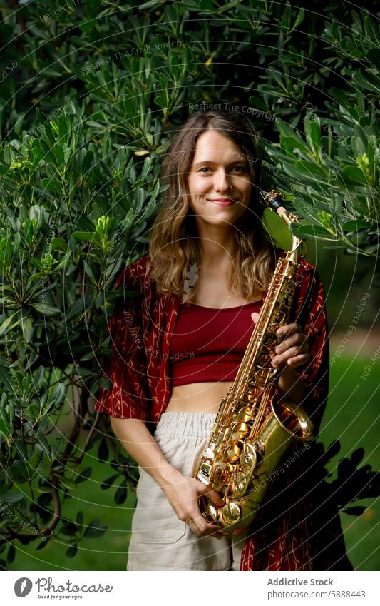 Frau mit Saxophon in einem üppigen Garten Musik Musiker grün Laubwerk Lächeln heiter freundlich in die Kamera schauen saxowoman Instrument Messing Jazz